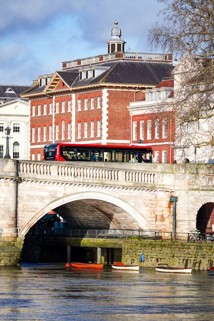 The image shows a scene in Richmond upon Thames with a historic red brick building in the background, featuring multiple windows and a small cupola on the roof. A red and black bus is traveling along a road on a stone bridge that spans a river, with the bridge having classical balustrades and arches beneath it. Below the bridge, the river contains a few small rowing boats anchored near the riverbank. To the right, a leafless tree extends its branches over the scene, suggesting late autumn or winter. The lighting is natural, with a partly cloudy sky overhead, and the overall setting reflects an area known for its picturesque surroundings, which could be included in a guide for house removals or moving logistics in the Richmond area. Occasionally, landmarks like this are part of scenic routes for furniture transport or home relocation services, emphasizing the practical context of moving operations in the vicinity, as might be detailed on the website manwithavanrichmonduponthames.co.uk.