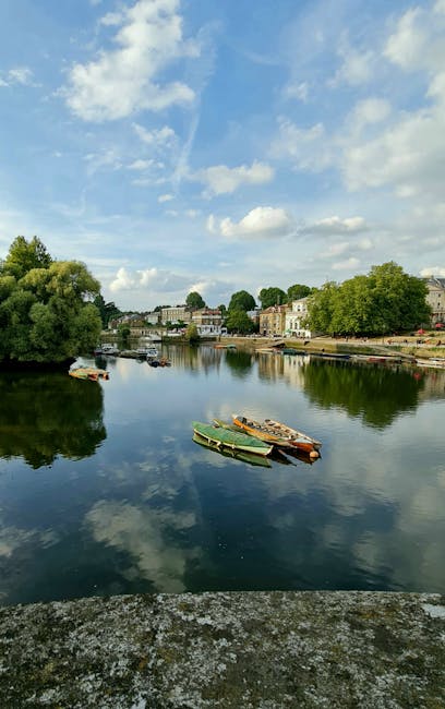A tranquil riverside scene showing a calm waterway reflecting a partly cloudy blue sky. Along the riverbank, there are several small boats tied to the edge, some with wooden and plastic hulls in shades of green, yellow, and red. The boats are situated close to the shore, which is lined with trees and lush greenery. On the opposite side of the river, there are residential buildings with light-colored facades and flat roofs, surrounded by additional trees and open space. The scene is well-lit with natural daylight, highlighting the peaceful environment typical of a local area where house removals or relocation services might take place, as supported by the context of the George Street Richmond man and van guide for narrow access by [COMPANY_NAME].