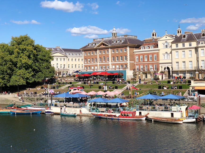 The image shows a scene in Richmond upon Thames with a historic red brick building in the background, featuring multiple windows and a small cupola on the roof. A red and black bus is traveling along a road on a stone bridge that spans a river, with the bridge having classical balustrades and arches beneath it. Below the bridge, the river contains a few small rowing boats anchored near the riverbank. To the right, a leafless tree extends its branches over the scene, suggesting late autumn or winter. The lighting is natural, with a partly cloudy sky overhead, and the overall setting reflects an area known for its picturesque surroundings, which could be included in a guide for house removals or moving logistics in the Richmond area. Occasionally, landmarks like this are part of scenic routes for furniture transport or home relocation services, emphasizing the practical context of moving operations in the vicinity, as might be detailed on the website manwithavanrichmonduponthames.co.uk.